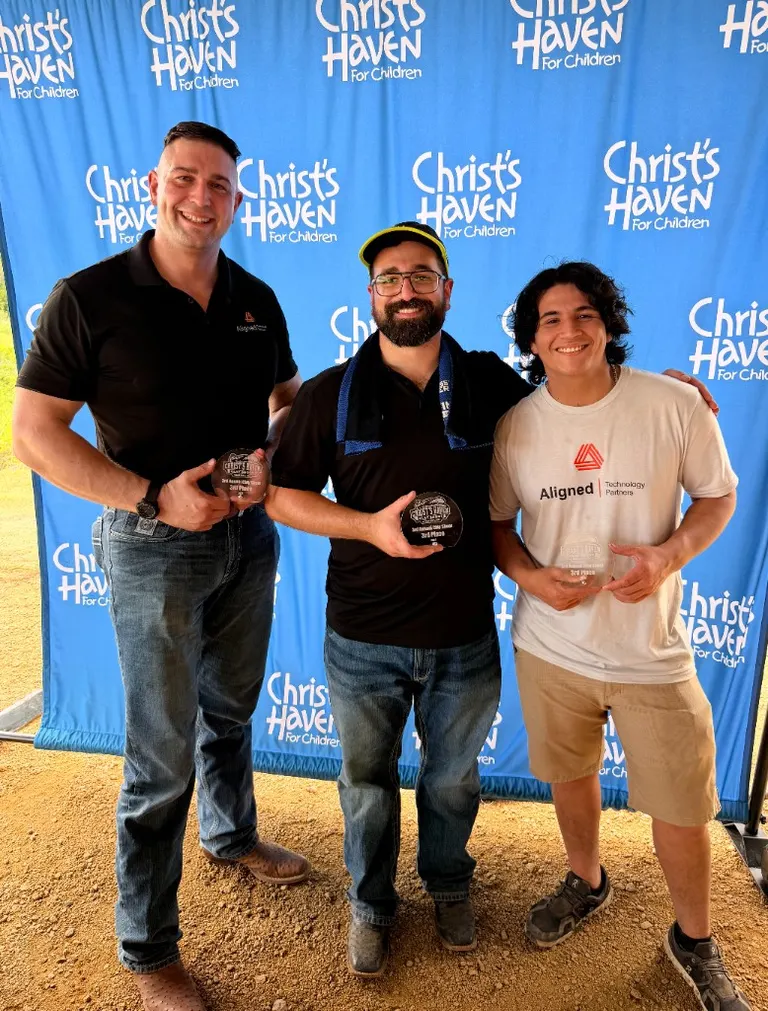 Three Aligned Technology Partners teammates holding third-place trophies in front of a Christ’s Haven for Children backdrop.